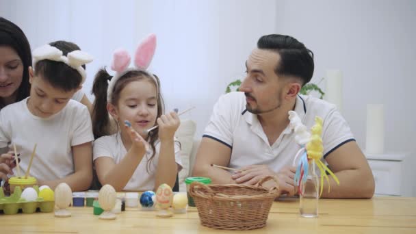 Les parents avec leurs adorables et mignons enfants, qui leur ressemblent vraiment, colorent les œufs de Pâques, assis à la table en bois, plein de décorations de Pâques. Petite fille peint sa paume dans une — Video