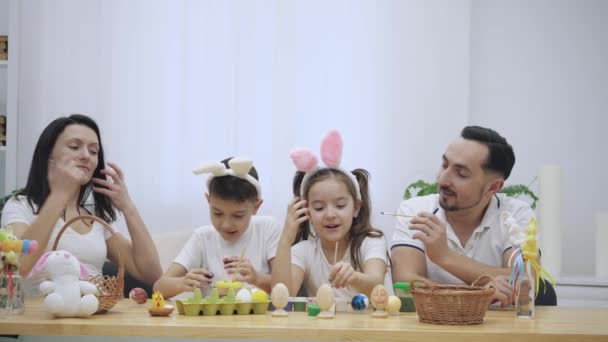 Γονείς με χαρούμενα και χαριτωμένα τα παιδιά τους, που είναι πολύ παρόμοια με τους, colourizing αυγά του Πάσχα, στο ξύλινο τραπέζι, γεμάτο διακοσμήσεις Πάσχας. Παίζει ο πατέρας με την κόρη του και — Αρχείο Βίντεο