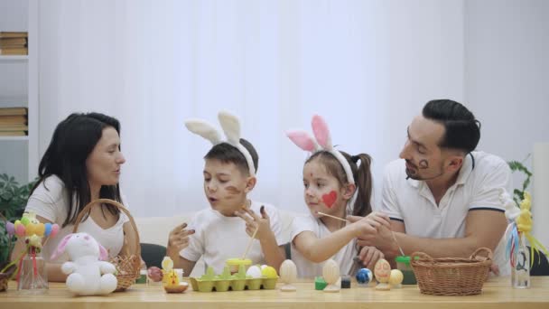 Ouders met hun jonge geitjes vrolijk en schattig, zijn colourizing elkaar, aan de houten tafel, vol met Pasen decoraties. Moeder is schilderen met gele verf op de rechterwang zonen, maar zoon is — Stockvideo