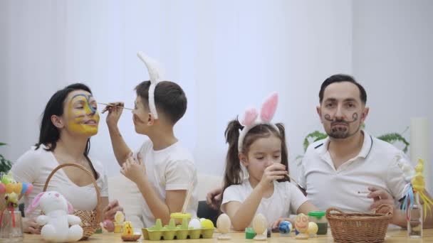 Eltern mit ihren flotten und kleinen Kindern bemalen sich gegenseitig, sitzen am Holztisch voller Osterdekorationen. — Stockvideo