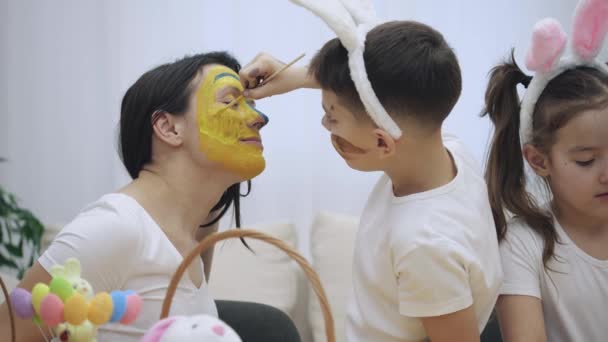 Pequeño niño lindo con orejas de conejito en la cabeza está pintando la cara de las madres en colores amarillo y azul. Concepto vacaciones de Pascua . — Vídeos de Stock