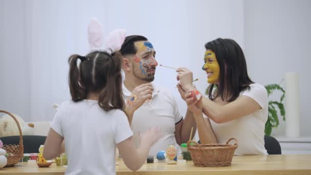 Das süße Paar färbt sich gegenseitig mit einem Osterpinsel ein. Plötzlich bemerkten sie, dass ihre Tochter sie ansah. Sie fangen an, Ostereier zu bemalen, wie es die ganze Zeit war. — Stockvideo