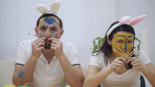 Linda y adorable pareja volvió a la infancia. El hombre sostiene dos pinceles en sus manos y una mujer sostiene cuatro pinceles en ella. Pareja con orejas de conejo. Se están burlando. — Vídeos de Stock