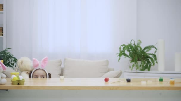 Kleine spielende Kinder mit Hasenohren auf dem Kopf verstecken sich unter dem Holztisch voller Osterdekorationen. Nettes Mädchen, dann schaut ihr Vater am Ende raus. — Stockvideo