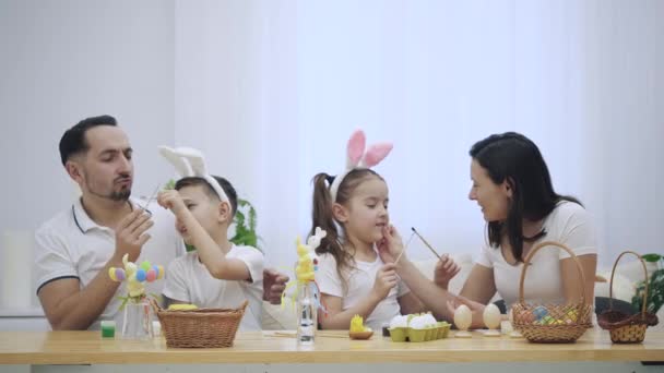 Glückliche und positive Familie: Mutter, Vater und zwei Kinder lächeln auf dem weißen Hintergrund. Sie spielen mit Pinseln. Mama gibt ihrer entzückenden Tochter einen kleinen Kuss. Osterferienkonzept — Stockvideo