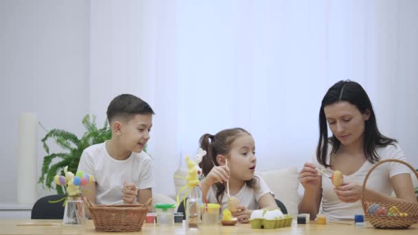 Madre está con sus hijos: hija e hijo, que están sentados en la mesa de vacaciones con una decoración de Pascua. La familia está haciendo artesanía, coloreando los huevos de Pascua . — Vídeos de Stock
