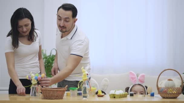 Des petits frères et sœurs se cachent sous une table, pleine de décorations de Pâques, tandis que leurs parents entrent et regardent, touchant les objets, qui sont situés sur la table. Les parents recherchent — Video