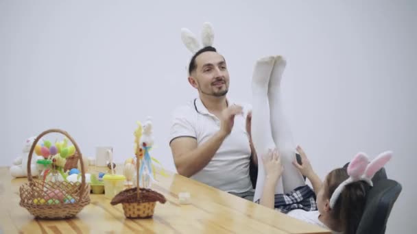 O pai está a brincar com a filha. Papai está brincando com filhas pequenas pernas vestidas de collants. A filha está a sorrir alegremente. Há uma mesa com decorações de Páscoa no vídeo . — Vídeo de Stock