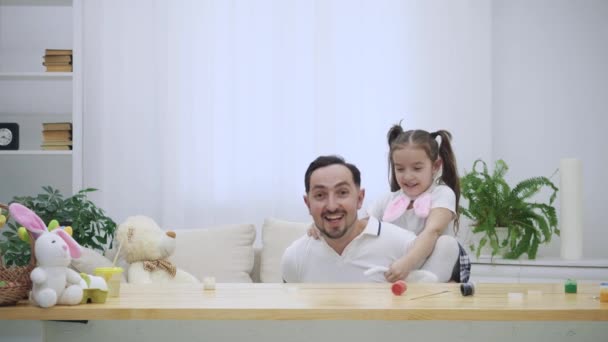 Filha feliz e alegre está sentada nos ombros de seus pais e dançando. Pai está de pé da mesa de madeira, onde existem algumas tintas coloridas. Menina está vestindo orelhas de coelho em — Vídeo de Stock