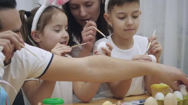 Los padres con sus adorables y lindos hijos, que son muy parecidos a ellos, están empezando a colorear los huevos de Pascua, sentados en la mesa de madera, llenos de decoraciones de Pascua . — Vídeo de stock