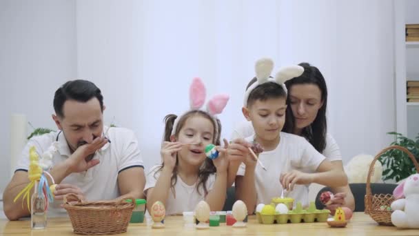 Los padres con sus adorables y lindos hijos, que son realmente similares a ellos, están coloreando los huevos de Pascua, sentados en la mesa de madera, llenos de decoraciones de Pascua. La familia se concentra en la actividad — Vídeo de stock