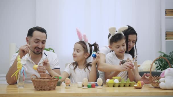 Os pais com seus filhos adoráveis e fofos, que são realmente semelhantes a eles, estão colorindo ovos de Páscoa, sentados à mesa de madeira, cheios de decorações de Páscoa. Família concentra-se na atividade — Vídeo de Stock