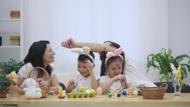 Os pais com seus filhos alegres e bonitos, estão se colorindo, sentados à mesa de madeira, cheios de decorações de Páscoa. O pai está a fazer manchas no nariz das esposas. Filho se junta e faz o — Vídeo de Stock