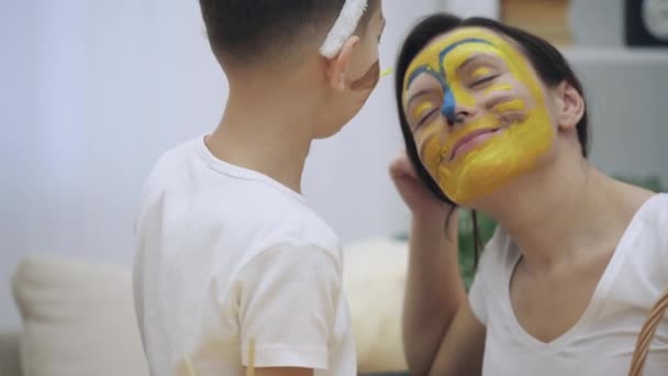 Pequeño niño lindo con orejas de conejito en la cabeza está pintando la cara de las madres en colores amarillo y azul. Concepto vacaciones de Pascua. Moomy sonríe sinceramente. . — Vídeo de stock