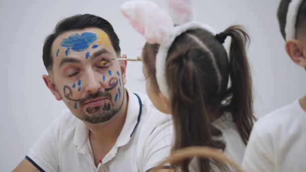 Little cute girl with bunny ears on her head is painting daddys face in yellow and blue colors. Concept Easter holiday. — Stock Video