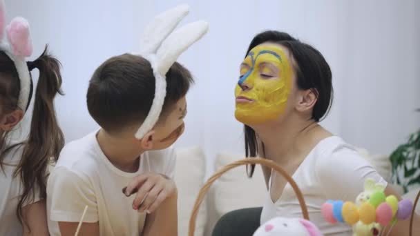 Little cute boy with bunny ears on his head has colorised mothers face in yellow and blue colors. Concept Easter holiday. Moomy and son are smiling sincerely. — Stock Video