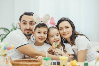Mutlu aile masada oturan tam og Paskalya malzemeleri, sarılma ve gülümseyerek. Komik Tavşan kulakları çocuklarda. Pigtails ile şirin kız renkli yumurta tutuyor.