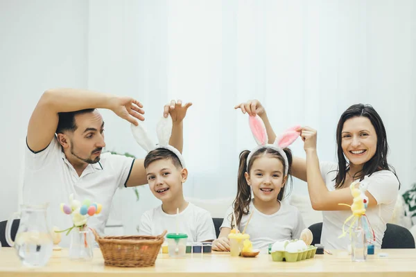 Funny lovely sister, brother and their parents playing around during the exciting process of painting eggs for Easter. Mother and father raising up the bunny ears on the heads of their children. — Stock Photo, Image