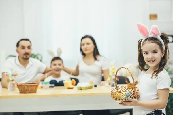 Hübsches kleines Mädchen mit Zöpfen und Hasenohren an der Spitze mit dem gelben Osterei in der Hand. Mutter und Bruder des Vaters sitzen am Tisch und bereiten sich auf Ostern vor. — Stockfoto