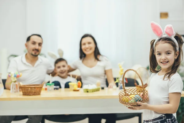 Dość mały dziewczyna z warkoczykami i uszy królika, stojąc na czele z koszem kolorowych jaj w jej ręce. Jej ojciec matka i brat siedział przy stole, przygotowanie do Wielkanocy. — Zdjęcie stockowe