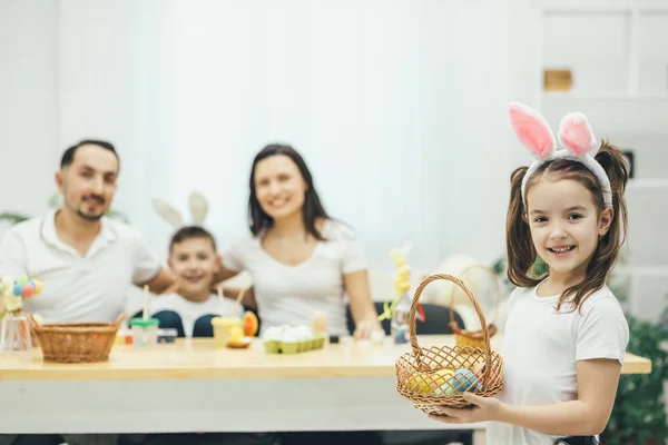 Dość mały dziewczyna z warkoczykami i uszy królika, stojąc na czele z koszem kolorowych jaj w jej ręce. Jej ojciec matka i brat siedział przy stole, przygotowanie do Wielkanocy. — Zdjęcie stockowe