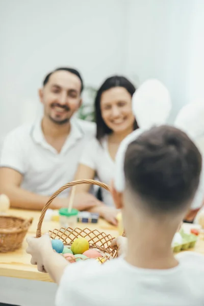 Little son giving his parents a basket of colored easter eggs. Mother and father sitting at the table, happy to get such beautiful present. — Stock Photo, Image