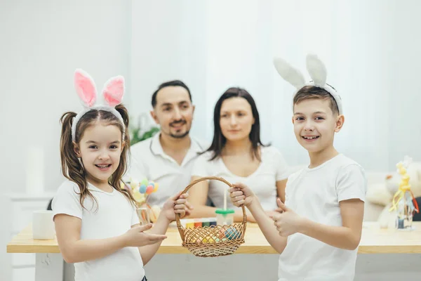 Niedliche kleine Kinder mit Hasenohren, die vorne stehen und einen Korb mit Ostereiern in der Hand halten. Eltern sitzen am Tisch auf dem verschwommenen Hintergrund. alle lächeln. — Stockfoto