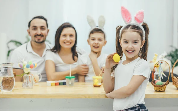 Dość mały dziewczyna z warkoczykami i uszy królika, stojąc na czele z żółtym Pisanka w jej dłoni. Jej ojciec matka i brat siedział przy stole, przygotowanie do Wielkanocy. — Zdjęcie stockowe