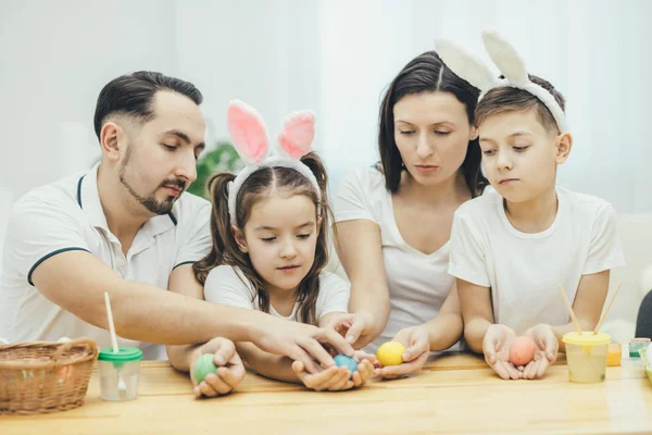 Inspirowane rodziny w białe koszulki razem siedział przy stole, trzymając kolorowe pisanki w ich rękach, patrząc na nich podnieceniu, wybierając, które jajko jest najlepszy. — Zdjęcie stockowe