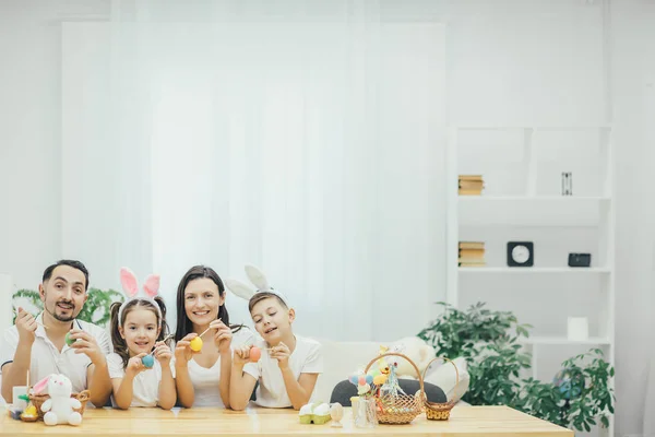Eine inspirierte Familie sitzt zusammen am Tisch, Bruder und Schwester in Hasenohren. Jeder bemalt verschiedene Ostereier mit unterschiedlichen Farben. — Stockfoto
