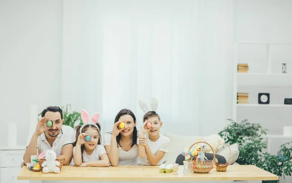 Glückliche Familie, Vater Mutter Sohn und Tochter verstecken eines ihrer Augen hinter bunten Ostereiern. Bruder und Schwester tragen Hasenohren. — Stockfoto