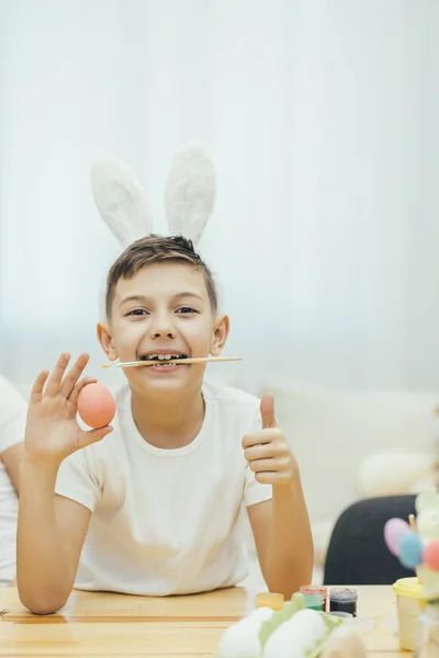 Pequeño niño lindo escondido mostrando huevo rojo a la cámara, y dando su pulgar hacia arriba, pincel en sus dientes . —  Fotos de Stock