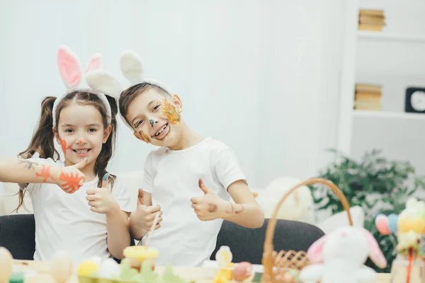 Pretty funny siblings brother and sisiter coloured their faces instead of easter eggs and now they are looking at the camera smiling and giving their thumbs up. — Stock Photo, Image