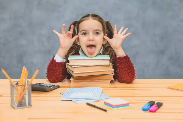 Una niña linda juguetona se está divirtiendo, confiando en libros gruesos sobre un fondo gris. Su cabello está hecho con arneses. Abrir la boca muestra la lengua . — Foto de Stock