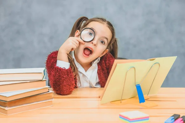 A playful little young little schoolgirl sits at the table reading a book. Her hair has two harnesses. During this, she looks at the camera through his lips opening his mouth. — Stock Photo, Image