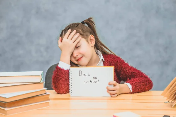 En liten skolflicka på en grå bakgrund. Håll papperet med texten tillbaka till skolan. Efter att ha låtit huvudet ner, flickan lägga sin hand på huvudet. — Stockfoto