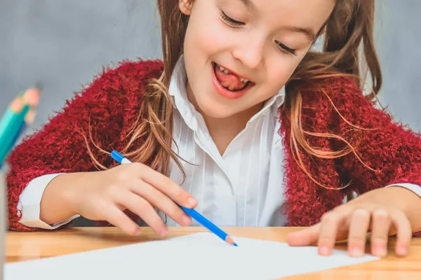 Close-up de uma menina bonita em um fundo cinza. Tomar um lápis azul gosta do processo de desenho. Na mesa há lápis cor de rosa . — Fotografia de Stock