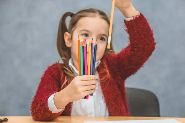 片手の女の子に色鉛筆のクローズアップコンセプト。黒い鉛筆を持って見る。灰色の背景。人形は鉛筆を見る。赤いジャケットを着て. — ストック写真