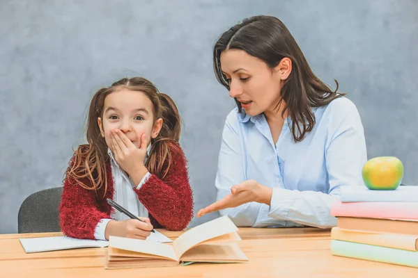 Mom helps me do my daughters homework. During this they are sitting on a gray background. A schoolgirl closed her mouth with her hand, a young woman shows her hand in a notebook.