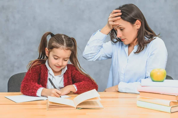 Genç anne ve kızı gri bir arka plan üzerinde oturuyor. Bu esnada anne, bir kızın ev ödevini nasıl yaptığına dikkat eder. Sürprizden elini alnına koyar.. — Stok fotoğraf