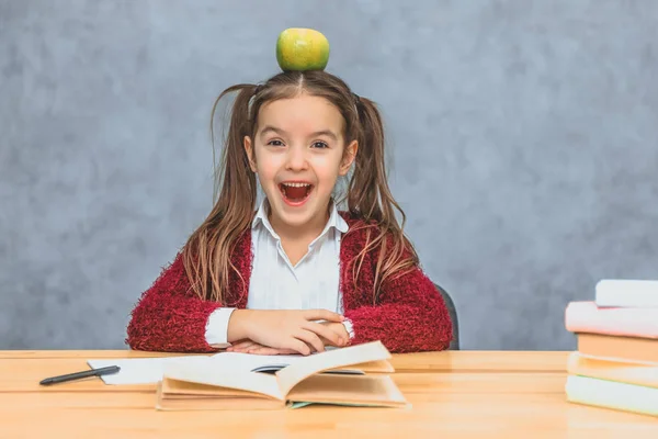 Portrait of cute clever girl with apple on her head. Sitting from a stack of books at the table, a copy of the space. Concept of education and development. — Stock Photo, Image