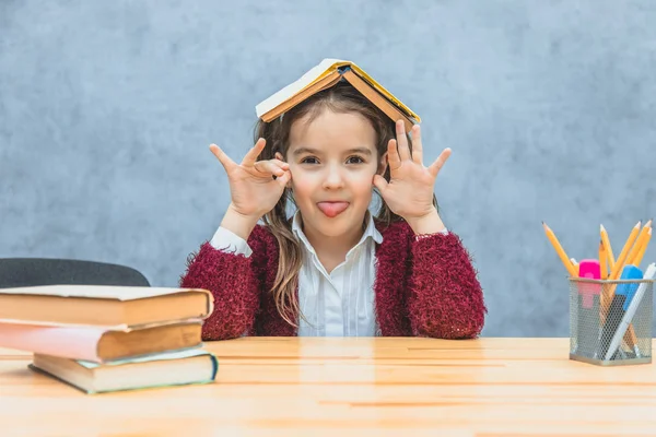 A cheerful girl sitting and holding a book over her head over a gray background. During this schoolgirl opened her mouth showing a tongue looking at the camera. — Stock Photo, Image