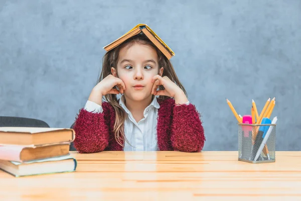 Jolie fille bouclée assise et tenant un livre au-dessus de sa tête sur un fond gris. Au cours de cette écolière prend ses yeux loin. Produits de papeterie sur la table . — Photo