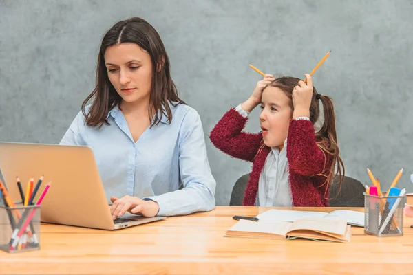 Córka i matka na szarym tle. W tym czasie dziewczyna stawia dwa żółte kredki na głowie, pokazując język. Mama Wygląda poważnie podczas pracy na laptopie. — Zdjęcie stockowe