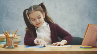 İyi bir kız öğrenci gri bir arka plan üzerinde masada oturur. Bu süre zarfında, kalem alır ve bir ev ödevi yazmaya başlar. Yavaş video.