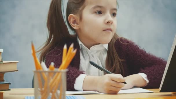 Ein braves Schulmädchen sitzt am Tisch vor grauem Hintergrund. Während dieses Schreibens lächeln die Hausaufgaben. Zeitlupe. — Stockvideo