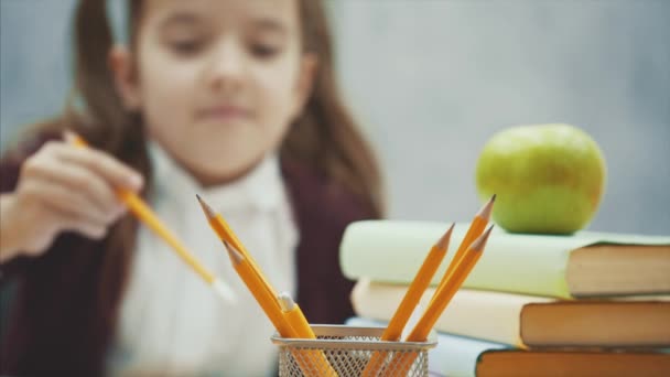 Nahaufnahme eines schönen Schulmädchens sitzt am Tisch auf grauem Hintergrund. nimmt dabei einen Bleistift von gelber Farbe. — Stockvideo