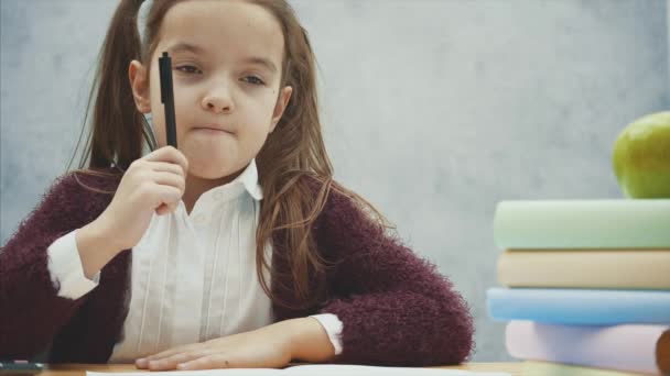 Közeli fel egy gyönyörű iskolás ül az asztalnál egy szürke háttér. Ez idő alatt, kezében a toll és a gondolkodás az írás. Gondosan feltalálta az írást. — Stock videók