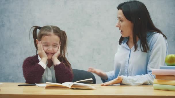 Uma menina em um fundo cinza senta-se à mesa e chora. Durante isso, minha mãe está brigando com sua filha. Conceito de relações . — Vídeo de Stock