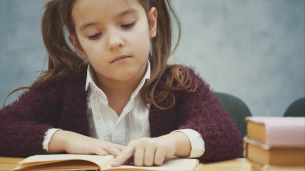 Linda niña sobre un fondo gris. Se sienta a la mesa y lee un libro . — Vídeos de Stock